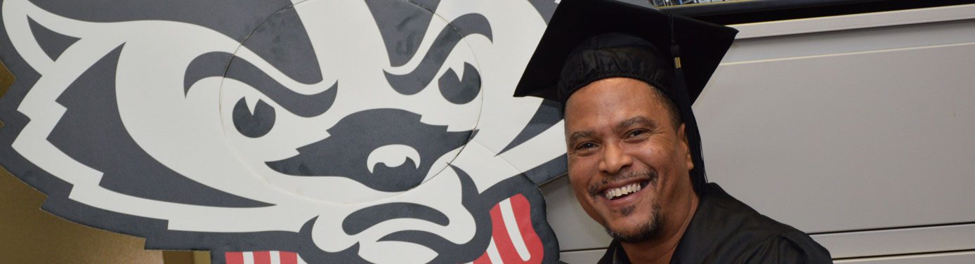 Happy Odyssey graduate Brian Benford poses next to Bucky Badger cardboard cutoff wearing cap and gown