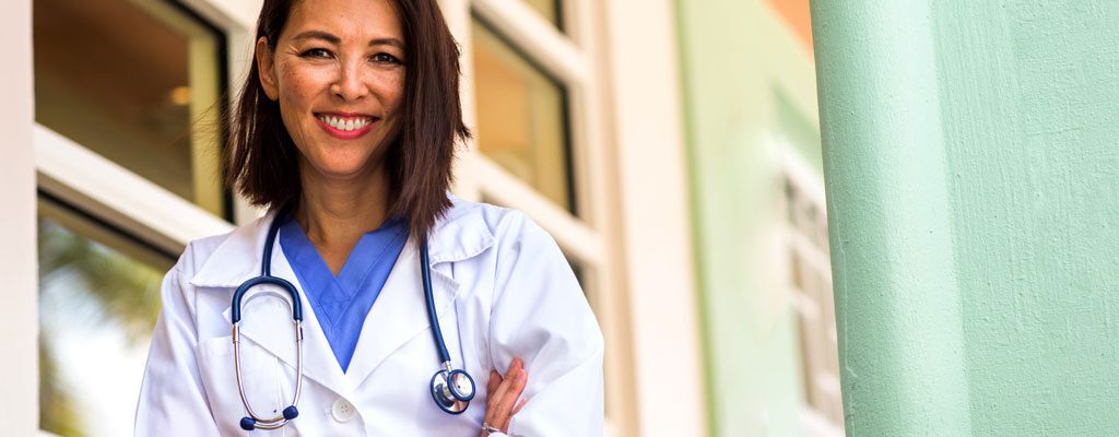 Smiling doctor wearing scrubs and stethoscope poses outside of building