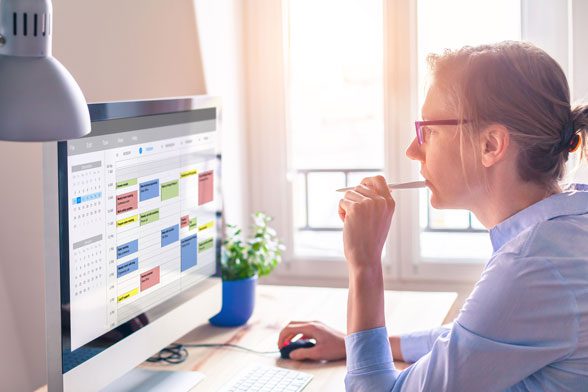 Woman sitting in front of computer with pencil in her mouth, considering her calendar