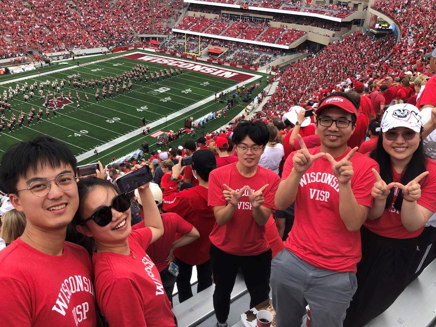 Visiting student alums face the camera at a Badger Football game and smile. Some flash the "W" with their fingers.