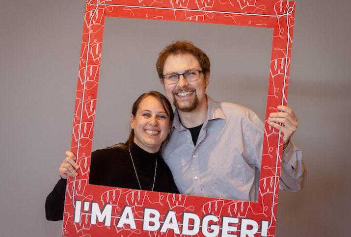 Tyler Johnston, a white man with brown hair and glasses, stands next to his wife, a white woman with black turtleneck. Both are standing, smiling, and hold an oversized red frame in front of them. The frame has the words I'm a Badger at the bottom.