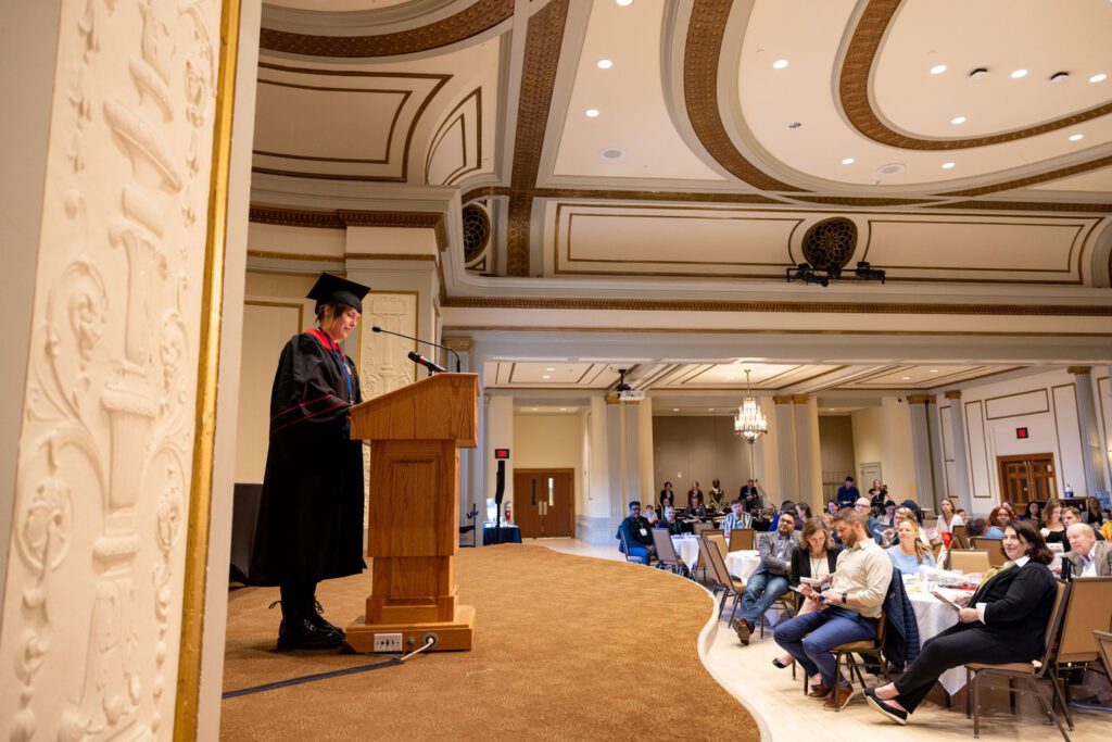 Student in graduation regalia stands at a podium presenting before a large audience of adult students and guests 