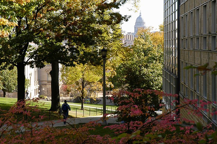 People walk up Bascom Hill as the tree foliage begins to change colors during autumn. At right is the Law School building. In the background is the Wisconsin State Capitol building dome.