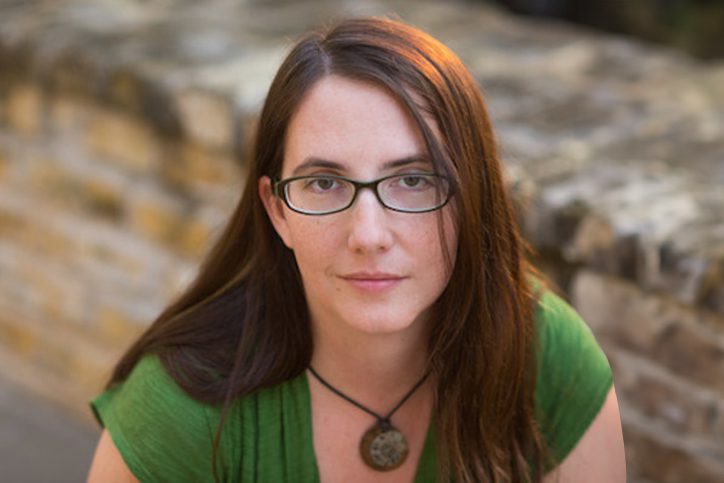 Megan Reilly, shown in an outdoor setting, is wearing a green v-neck shirt, a glasses and pendant necklace. She has long reddish-brown hair.