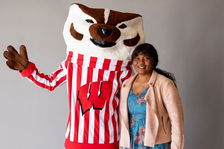 Zalissa Zongo Kafando stands directly next to Bucky Badger, who is wearing a striped shirt with the Wisconsin "W" on the front and waving. Zalissa is smiling and has dark hair pulled back in a low ponytail. She is wearing a blue floral dress with a peach-colored jacket.