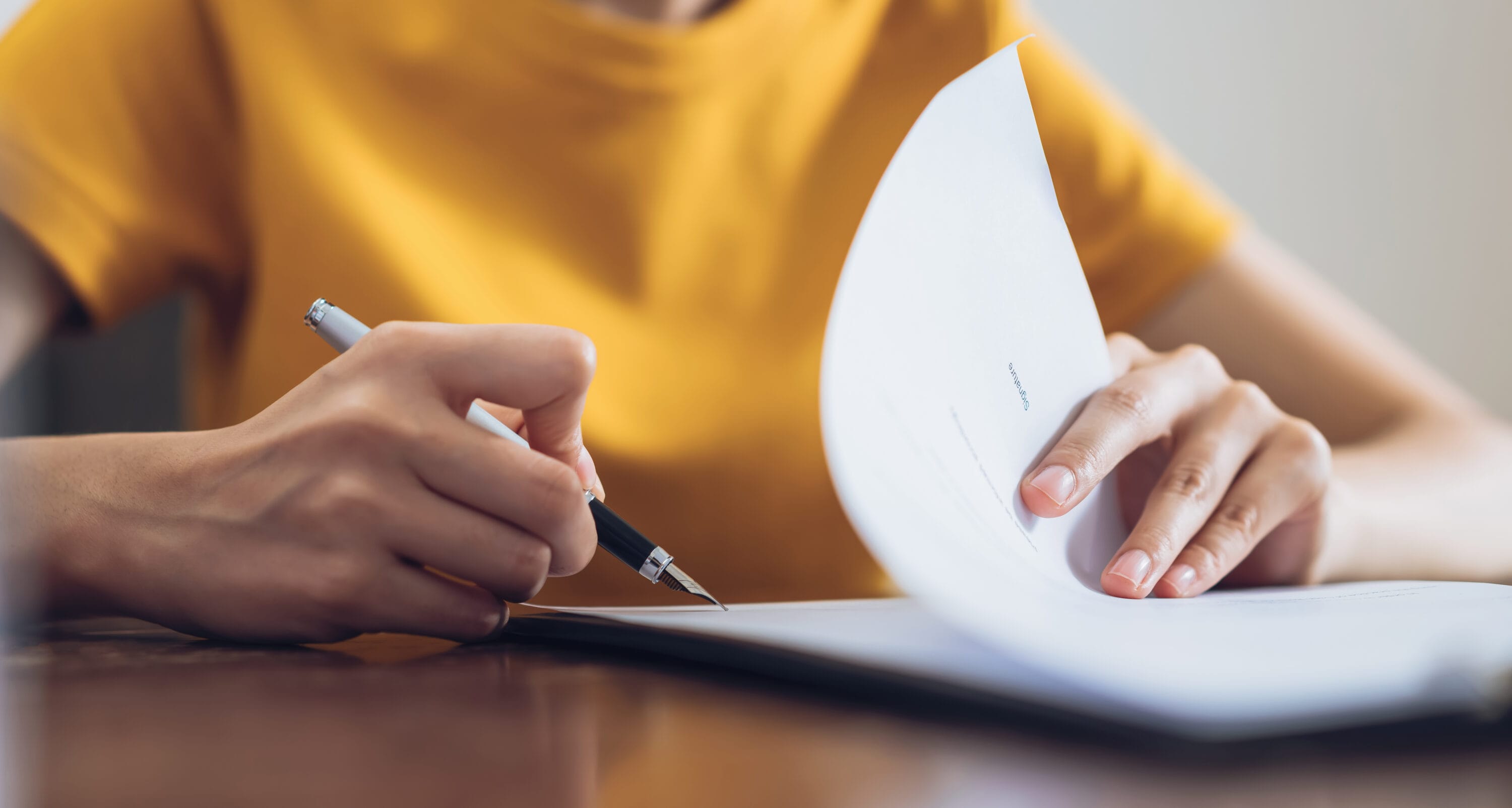 Person in yellow shirt signing document and hand holding pen putting signature at paper.