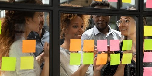 A group of people huddled around a glass board with sticky notes.