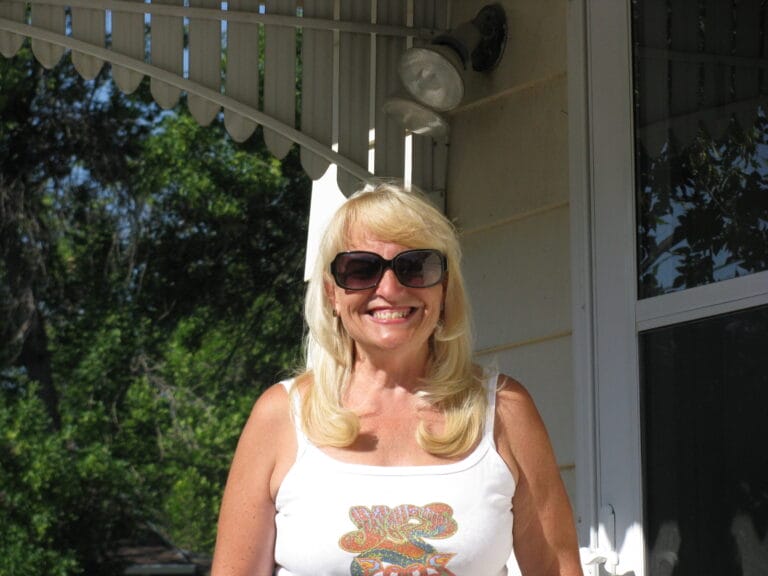 Jeannette Eichholz stands in a tank top and sunglasses on her front porch on a sunny day
